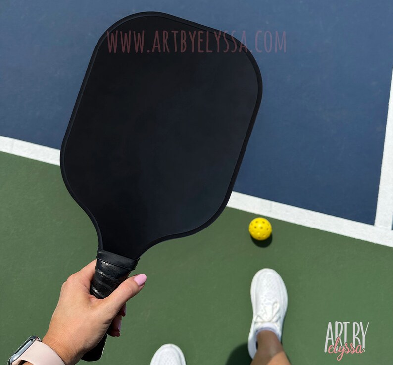 May include: A black pickleball paddle with a black grip. The paddle is held by a person on a pickleball court. A yellow pickleball is on the court.