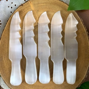 May include: Five white selenite crystal wands with a serrated edge. The wands are arranged in a row on a wooden surface.