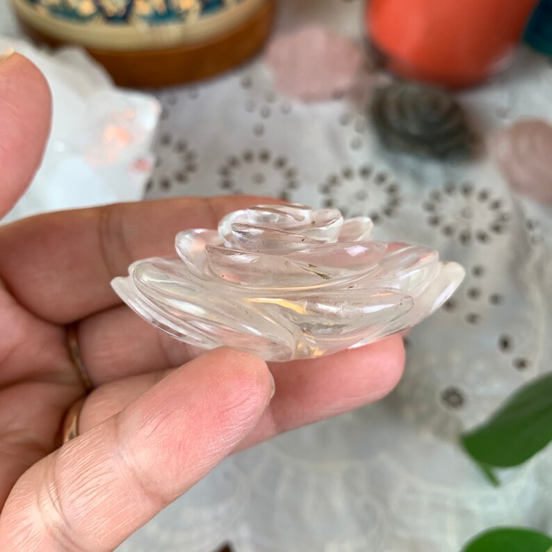 May include: A clear quartz crystal carved into the shape of a rose. The rose is made up of multiple layers of petals, each with a slightly different shape and size. The crystal is smooth and polished, and it has a delicate, feminine appearance.
