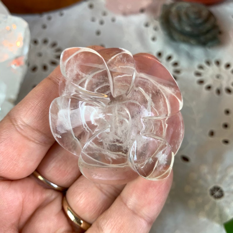 May include: A clear quartz crystal carved into the shape of a flower. The crystal is translucent and has a slightly cloudy appearance. The flower has multiple layers of petals.