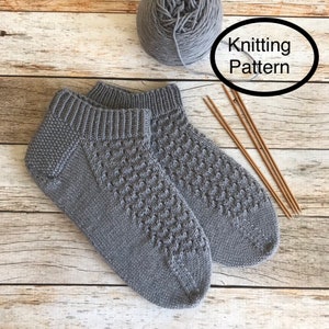 May include: A pair of gray knitted socks with a textured pattern. The socks are laid out on a wooden surface with knitting needles and a ball of gray yarn.