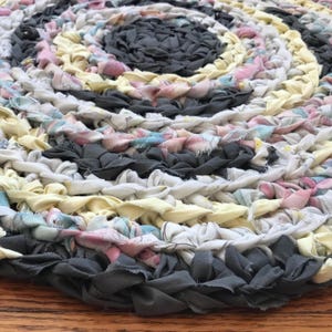 May include: Circular braided rug in a spiral pattern. The rug features a mix of fabric strips in various colors, including gray, yellow, pink, and white. The rug is handmade and rests on a wooden surface.