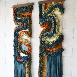May include: Two vertical wall hangings with textured, abstract designs. Each features a wooden rod at the top, with a mix of blue, orange, green, and cream-colored yarn. The bottom has a fringe of teal yarn. The background is a white brick wall.