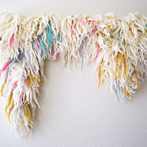May include: A white fluffy garland with colorful strands of yarn hanging down. The garland is hung on a wall with a metal rod.