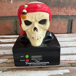 Könnte beinhalten: Ein schädelartiger Wecker mit rotem Bandana und schwarzem Kragen. Die Uhr hat eine Digitalanzeige und den Text "Pirates of the Caribbean: At World's End". Er verfügt über grüne und rote Tasten mit der Aufschrift "SET" und "MODE".