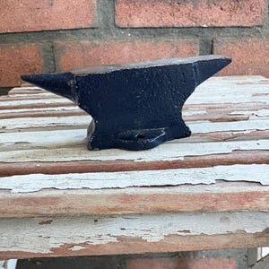 May include: A small, black anvil with a flat top and a pointed horn. The anvil is made of metal and sits on a weathered wooden surface. The background features a red brick wall.