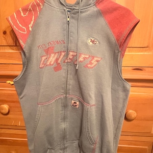 Op de afbeelding: Grijze mouwloze hoodie met rode accenten op de schouders. De voorkant toont de tekst "CHIEFS" in het rood, samen met een teamlogo. De hoodie heeft een voorzak en een capuchon met trekkoord. Afmetingen in cm.