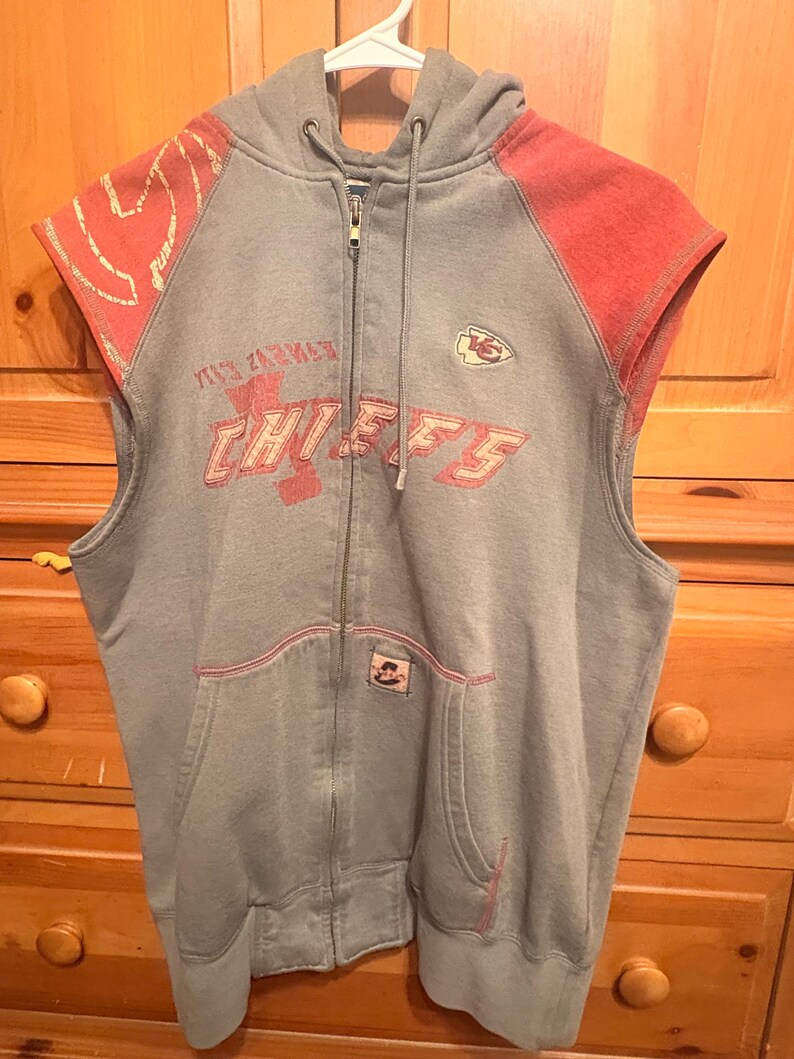 Op de afbeelding: Grijze mouwloze hoodie met rode accenten op de schouders. De voorkant toont de tekst "CHIEFS" in het rood, samen met een teamlogo. De hoodie heeft een voorzak en een capuchon met trekkoord. Afmetingen in cm.