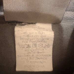 Op de afbeelding: Close-up van een grijs hoodie-label met wasvoorschriften en materiaalsamenstelling. Het label vermeldt "55% POLYESTER, 45% KATOEN" en "MADE IN CHINA." De maat is M.
