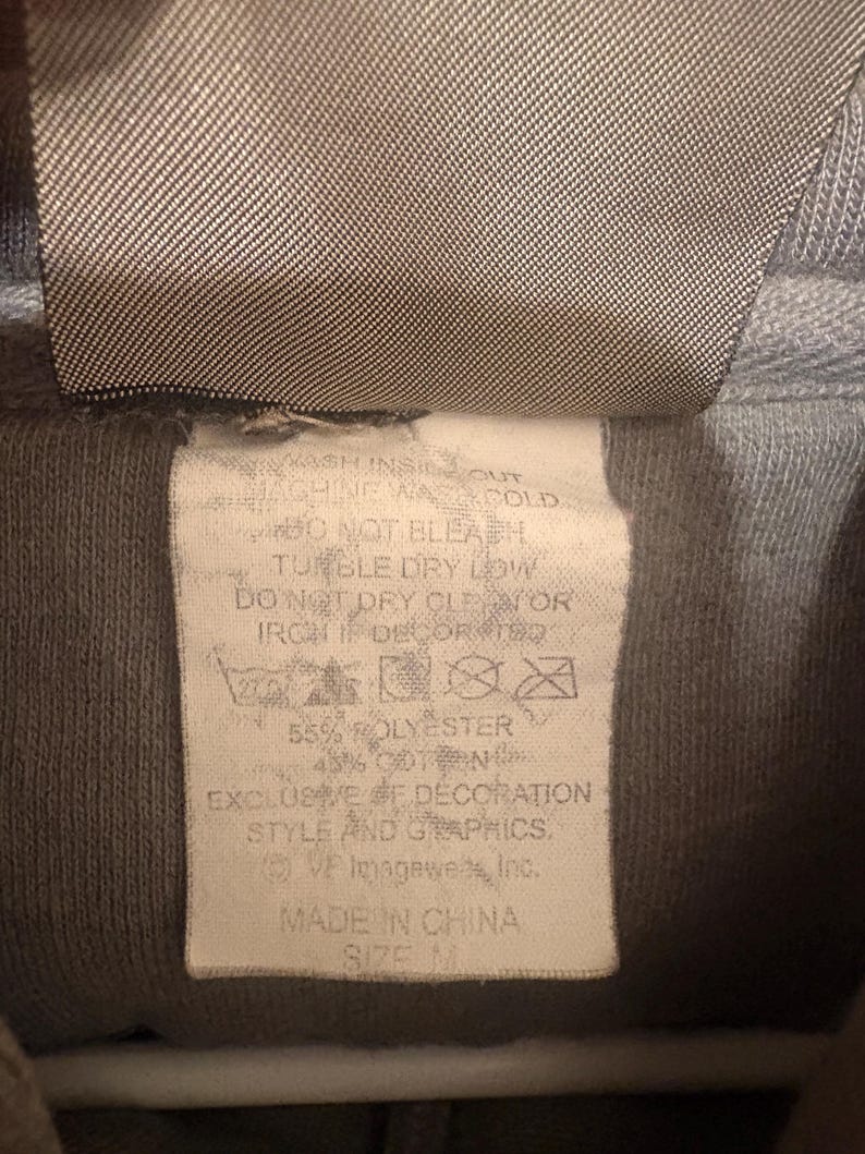 Op de afbeelding: Close-up van een grijs hoodie-label met wasvoorschriften en materiaalsamenstelling. Het label vermeldt "55% POLYESTER, 45% KATOEN" en "MADE IN CHINA." De maat is M.