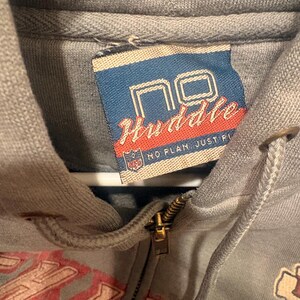 Op de afbeelding: Close-up van een grijze hoodie met ritssluiting en trekkoord. Het label leest "NO Huddle" in rode en witte tekst op een blauwe achtergrond. De rits is goudkleurig.