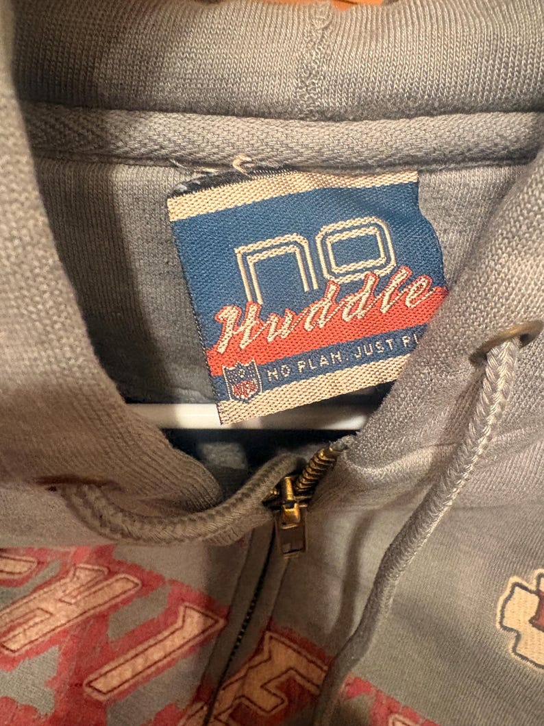 Op de afbeelding: Close-up van een grijze hoodie met ritssluiting en trekkoord. Het label leest "NO Huddle" in rode en witte tekst op een blauwe achtergrond. De rits is goudkleurig.