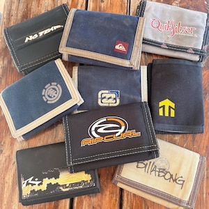 May include: A collection of various canvas wallets in different colors, including black, navy blue, and tan. Several wallets feature brand logos such as Rip Curl, Quiksilver, and Billabong. The wallets are arranged on a wooden surface.
