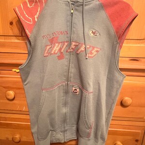 Op de afbeelding: Grijze mouwloze hoodie met rode accenten. De voorkant toont de tekst "CHIEFS" in het rood, samen met een teamlogo. De hoodie heeft een capuchon met trekkoord en een voorzak.