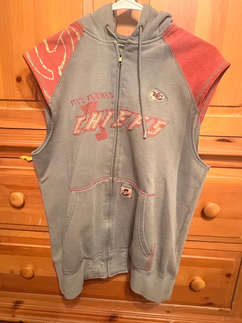 Op de afbeelding: Grijze mouwloze hoodie met rode accenten. De voorkant toont de tekst "CHIEFS" in het rood, samen met een teamlogo. De hoodie heeft een capuchon met trekkoord en een voorzak.