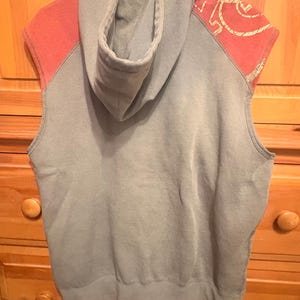 Op de afbeelding: Een mouwloze hoodie in een gedempte grijze kleur, met koraalkleurige schouderaccenten met een goudkleurig ontwerp. De hoodie heeft een capuchon en hangt aan een witte hanger.