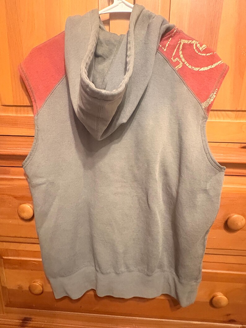 Op de afbeelding: Een mouwloze hoodie in een gedempte grijze kleur, met koraalkleurige schouderaccenten met een goudkleurig ontwerp. De hoodie heeft een capuchon en hangt aan een witte hanger.