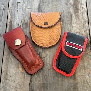 May include: Three small pouches in the image. One is brown leather, one is tan with a snakeskin pattern, and the third is black with red trim and the word "Craftsman". All are closed with a snap or flap.