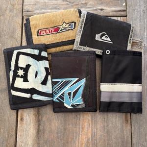 May include: A collection of five wallets in various colors and designs. One is corduroy with "Rusty" text, another black with a white logo, and a third with a white and black graphic. The others have blue and white designs.