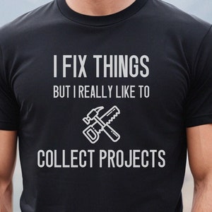 May include: Black t-shirt with white text that reads "I fix things but I really like to collect projects". A hammer and saw are crossed over the text.