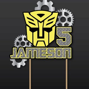 Pode incluir: Topper de bolo amarelo e preto com um rosto de robô, o número 5 e o nome "Jameson". O design inclui engrenagens prateadas em um fundo preto, com dois palitos de madeira para inserir.