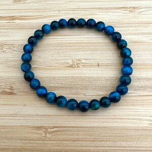 May include: A beaded bracelet featuring round, polished beads in shades of blue and teal. The bracelet is strung on an elastic band and is displayed on a light brown wooden surface. The beads have a marbled appearance.