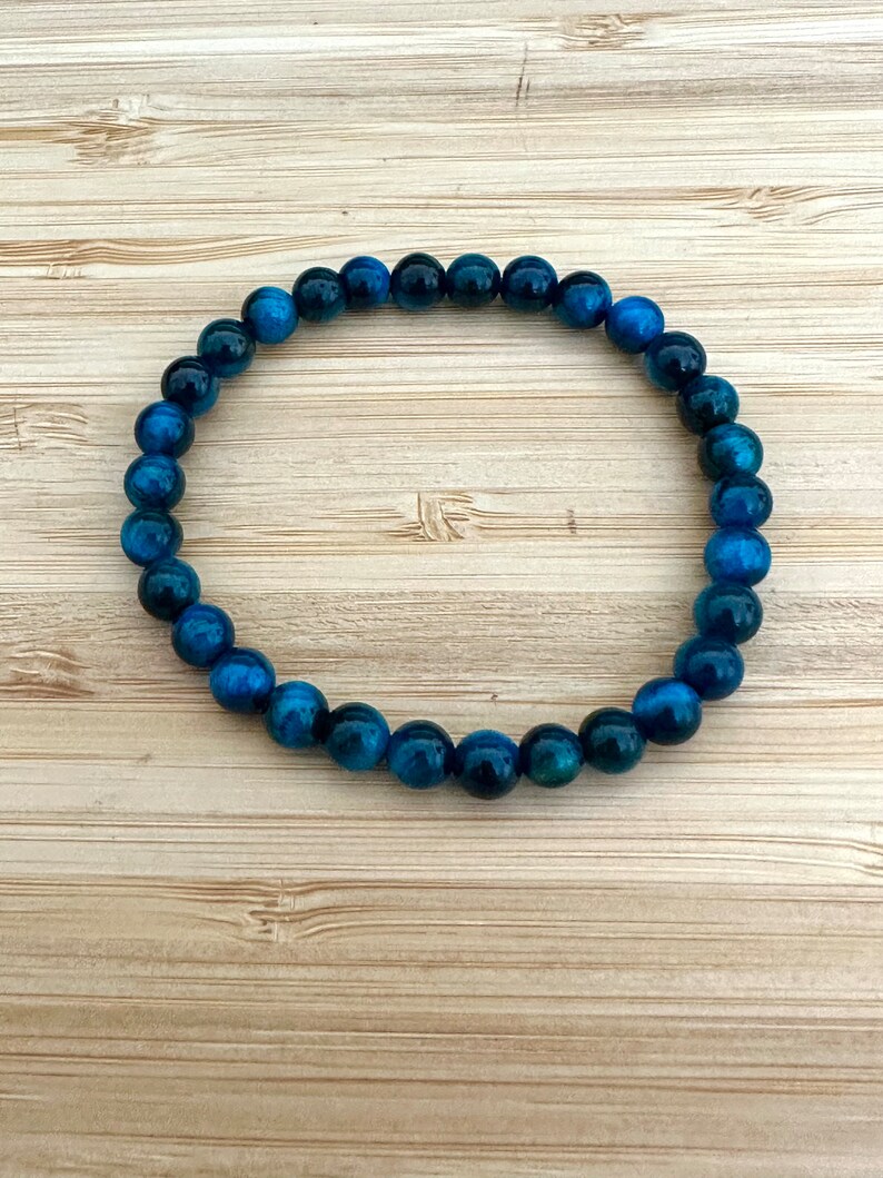 May include: A beaded bracelet featuring round, polished beads in shades of blue and teal. The bracelet is strung on an elastic band and is displayed on a light brown wooden surface. The beads have a marbled appearance.