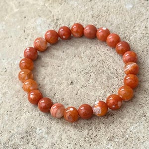 Op de afbeelding: Een cirkelvormige elastische armband bestaande uit gladde, ronde kralen in verschillende tinten oranje en roodachtig oranje. De kralen vertonen natuurlijke semi-transparante kwaliteiten met subtiele witte insluitingen en gemarmerde patronen.