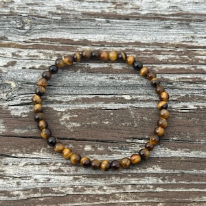 May include: A bracelet made of tiger's eye beads. The bracelet features a mix of brown, gold, and black beads. The bracelet is shown on a weathered wooden surface. The beads are approximately 0.8 cm in diameter.