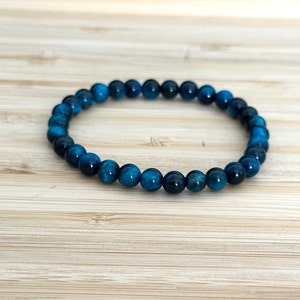 May include: A blue tiger eye beaded bracelet. The bracelet is made of small, round, polished beads in shades of deep blue and teal. The beads are strung together to form a flexible band. The bracelet is displayed on a light brown wooden surface.