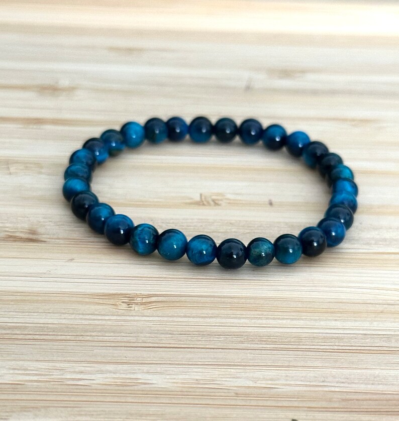 May include: A blue tiger eye beaded bracelet. The bracelet is made of small, round, polished beads in shades of deep blue and teal. The beads are strung together to form a flexible band. The bracelet is displayed on a light brown wooden surface.