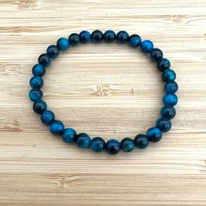 May include: A blue tiger eye beaded bracelet. The bracelet features round, polished beads in shades of deep blue and teal. The beads are strung together to form a circular shape, resting on a light wood grain surface.