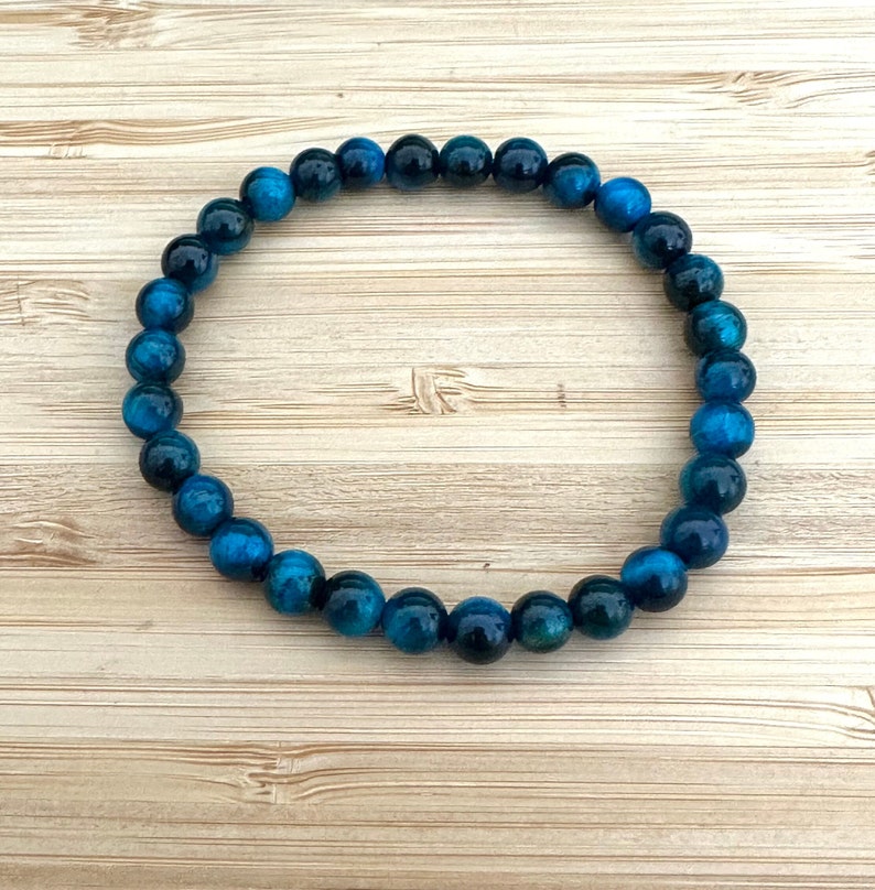 May include: A blue tiger eye beaded bracelet. The bracelet features round, polished beads in shades of deep blue and teal. The beads are strung together to form a circular shape, resting on a light wood grain surface.