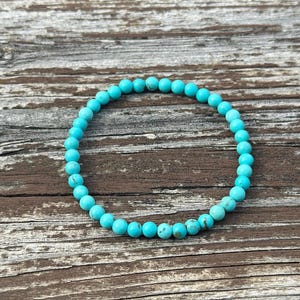May include: A turquoise beaded bracelet. The bracelet is made of small, round, light blue beads. The beads are strung together to form a circular shape. The bracelet is displayed on a weathered, brown wooden surface.