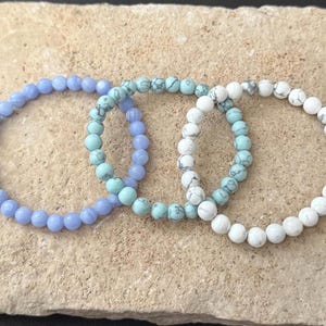 May include: Three stretch bracelets made of round beads. One bracelet features solid light blue beads, the second consists of turquoise beads with dark veining, and the third is composed of white beads with grey veining arranged in a circular pattern.