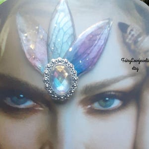 May include: A headpiece featuring a central oval iridescent cabochon set in a silver-toned metal frame, flanked by three translucent, wing-shaped accents in shades of blue, purple, and pink. The text FairyLovejewels etsy and Always purchase where you see my name on photo are visible.