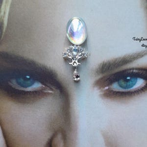 May include: A silver-toned bindi featuring an iridescent oval cabochon at the top, a central tree-like metal structure adorned with clear crystals, and a small dangling teardrop-shaped crystal at the bottom, displayed on a printed portrait.