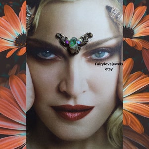 May include: A close-up of a woman's face with a small, ornate, green and purple gemstone forehead piece. The piece is made of metal and has a floral design. The text "Fairylovejewels etsy" is visible in the lower right corner of the image.
