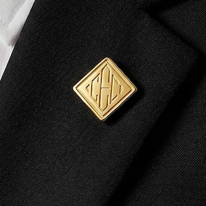 Pode incluir: Um alfinete de lapela dourado em forma de diamante com um design gravado, preso a um casaco preto. O alfinete está posicionado na lapela, perto de uma camisa branca e uma gravata preta. O design do alfinete parece ser iniciais ou um monograma.