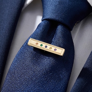 May include: A gold-coloured tie bar with a rectangular design, adorned with a sequence of colourful gemstones. The tie bar is set against a navy blue tie and a white shirt, creating a classic and elegant appearance. Gemstones include orange, green, blue, yellow, and purple.
