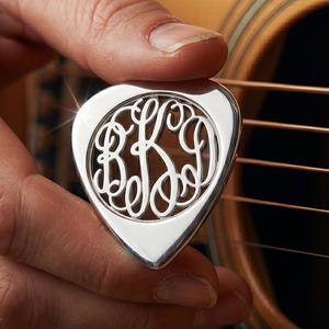 Könnte beinhalten: Ein poliertes silbernes Gitarrenplektrum mit einem kreisförmigen Ausschnittdesign, das die ineinander verschlungenen Monogrammbuchstaben B, K und D in einer dekorativen Schreibschrift zeigt. Die Metalloberfläche reflektiert das Licht.