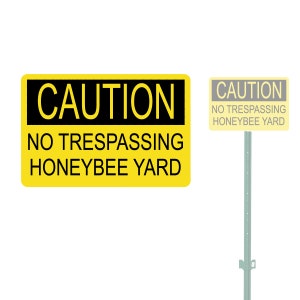 May include: Yellow caution sign with black text that reads "CAUTION NO TRESPASSING HONEYBEE YARD". A smaller yellow sign with black text that reads "CAUTION NO TRESPASSING HONEYBEE YARD" is mounted on a metal post.