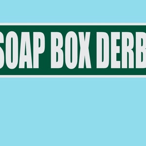 May include: A green street sign with white text that reads "SOAP BOX DERBY ST". The sign is attached to a metal pole.