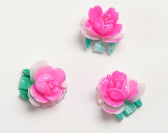 May include: Three small, decorative flower embellishments. Each flower has bright pink petals, white accents, and green leaves. The flowers are arranged on a white background.