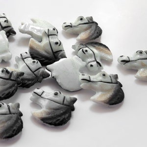 Puede incluir: Una colecci&oacute;n de peque&ntilde;as figuras de cabeza de caballo talladas. Las figuras son principalmente blancas con detalles grises y marrones, y presentan bridas detalladas. Las cabezas de caballo est&aacute;n dispersas sobre una superficie blanca.