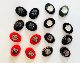 Puede incluir: Una colección de dieciséis botones de forma ovalada dispuestos en filas. Los botones presentan motivos de rosas en relieve en varios colores, incluidos negro, blanco, rojo y oro metálico, sobre bases ovaladas negras o rojas.
