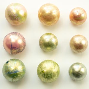Op de afbeelding: Negen iriserende glazen kralen in verschillende kleuren, waaronder goud, roze, groen en zilver. De kralen hebben een textuuroppervlak en een bolvorm. De kralen zijn gerangschikt op een witte achtergrond.