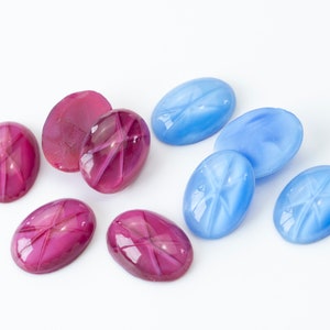 Könnte beinhalten: Sortiment ovaler Cabochon-Edelsteine in Rosatönen und Blau. Die Steine haben ein sternförmiges Muster auf ihrer Oberfläche.
