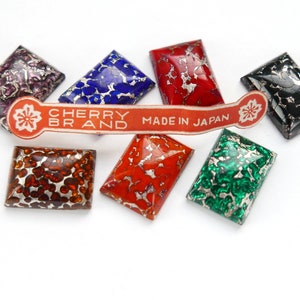 Puede incluir: Seis azulejos rectangulares, iridiscentes y decorativos con destellos plateados. Los azulejos son de varios colores, incluyendo rojo, naranja, verde, azul, negro y morado. Una etiqueta roja con el texto "CHERRY BRAND MADE IN JAPAN" está delante de los azulejos.