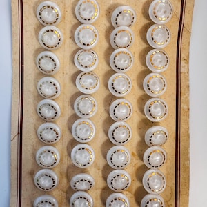 Könnte beinhalten: Eine Sammlung weißer Knöpfe mit einem goldfarbenen kreisförmigen Design. Die Knöpfe sind in Reihen auf einem Vintage-Kartonrücken mit rotem Rand angeordnet. Die Knöpfe haben ein perlmuttartiges Aussehen.